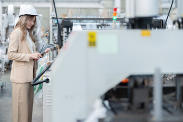 Asian female engineer uses her hands to press buttons on the control panel of a plastic and steel production machine in a large industrial factory. Wear a hard hat, have a laptop, radio, suit.