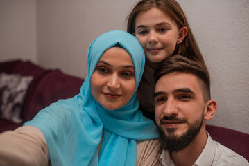 Two Arab parents and their daughter are sitting on the sofa while taking selfies together at home