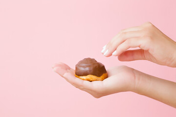 Young adult woman hand holding baked soft bun with chocolate glaze on light pink wall background....