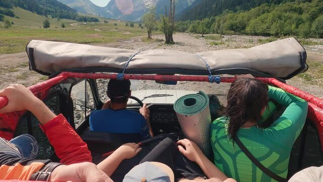 SUV with tourists driving on gravel stony in the mountains. First-person view of saloon with people and driver. Ahead are high mountains with snow on the tops. Off-road journey
