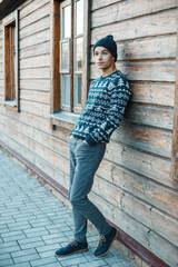 handsome young man model with a knitted hat in a fashion knitted sweater with a print stands near a wooden old rural house