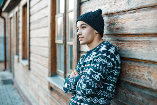 stylish handsome young man with a knitted hat in a vintage sweater with a pattern stands near a wooden rural house