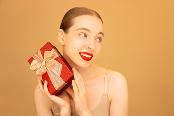 Portrait of cute girl with red gift box on peach isolated background. Happy beautiful Caucasian woman with red lipstick. Ads for getting a gift. Valentine's Day concept. International Womens-day