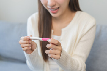 Happy thankful asian young woman hand holding pregnancy test after result with positive, surprised glad girl sitting on couch living room at home. Pregnant of ovulation fertility, motherhood concept.