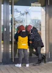 A small, sweet girl and boy look into the large glass door with great interest. Back view