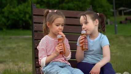 Little happy girls sit on bench fun drink apple juice from bottles with straws in summer park. Children leisure outdoors.