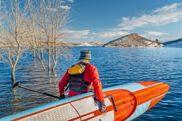 male paddler wearing a drysuit, life jacket and safety leash  is starting workout on a long racing stand up paddleboard on a mountain lake in Colorado - Horsetooth Reservoir in winter conditions