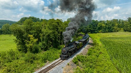 An Aerial View of a Classic Black Narrow Gauge Steam Train Chugging Along Tracks Surrounded By Lush Greenery And Emitting Thick Smoke Into The Clear Sky. - Powered by Adobe