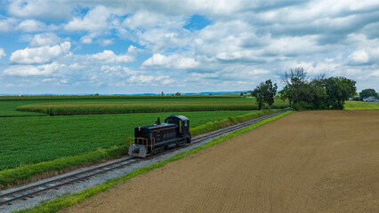 Fototapeta na wymiar Single Locomotive Traversing Along Railway Tracks Bordering A Plowed Field And Green Crops Under A Vast Sky With Puffy Clouds.