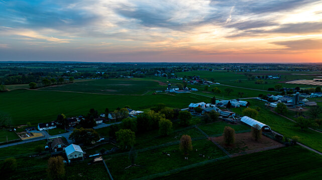 Sunset Casts A Warm Glow Over A Rural Farmland Tapestry With Homes, Barns, And Lush Fields Under A Dynamic Sky.