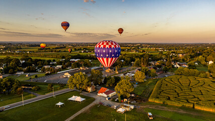 Patriotic-Themed Hot Air Balloon Dominates The Foreground With Others Dotting The Sky Above A Serene Rural Town As The Sun Sets.