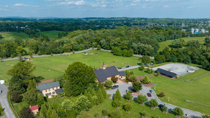 Fototapeta na wymiar Aerial View Of A Rural Landscape With Various Buildings, Including A Church, Scattered Across Rolling Green Fields Surrounded By Trees, With A Town In The Background Under A Wide Blue Sky.