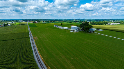 Expansive Aerial Perspective Of Multiple Farm Buildings Nestled Among Patchwork Fields With A Railway And Road Running Parallel Surrounded By The Verdant Countryside Under A Cloud-Dappled Sky.