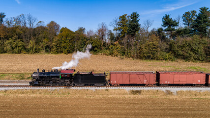 Fototapeta premium Vintage Steam Locomotive Pulling Red Freight Cars Along Tracks Adjacent To Golden Fields And Autumn Trees Under A Clear Blue Sky.