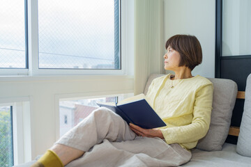 窓辺で読書しながら外を見るシニア女性