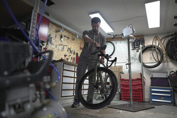 A man is repairing a bicycle in a garage, working on the tire and wheel rim. The building has a...