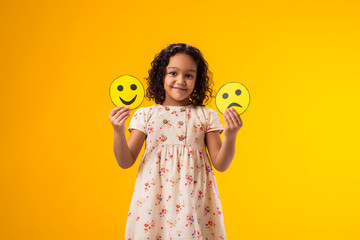 Smiling kid girl holding sad and happy emoticons in hands. Mental health, psychology and children's emotions concept - obrazy, fototapety, plakaty