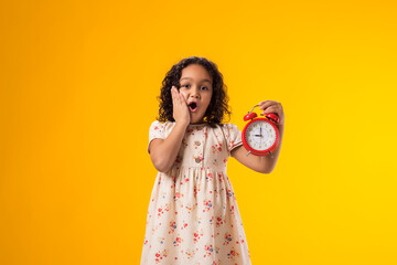 Surprised kid girl holding an alarm clock in hand. The concept of education, school, deadlines,...
