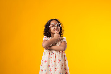 Thoughtful kid girl with glasses looking at camera over yellow background. Childhood and knowledge concept