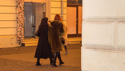 Two elegant women wearing coats walking at night on a lighted street in the city square. Evening...