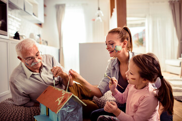 Family with grandfather, mother, and child painting a birdhouse together at home