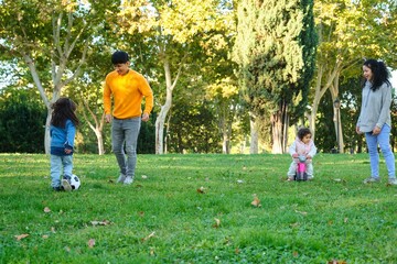 Fototapeta premium Hispanic family with two children playing soccer and with balance bike in a park. Latin family.