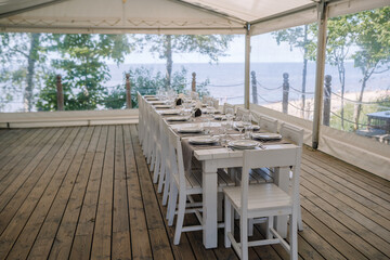 Jurmala, Latvia - july 25, 2023 - Outdoor dining setup under a tent on a wooden deck with a long table set for a meal, overlooking a beach and the sea.