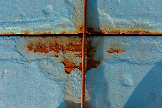 Superficie metalizada pintada de azul con zonas oxidadas y cuerda oxidada atraves&aacute;ndola en vertical.