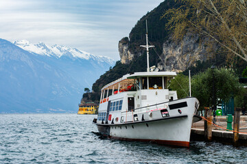 Waterfront view of the lake Garda with snowy mountains on the background and a boat in Riva del Garda, Trentino Alto Adige region, Italy. - obrazy, fototapety, plakaty