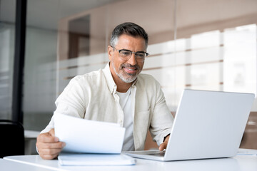 Smiling Latin or Indian male business man accountant analyst holding documents, work at laptop...