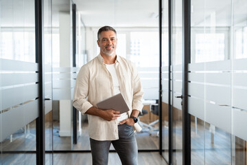 Portrait Latin Hispanic mature adult professional business man, smiling Indian senior businessman CEO walking and holding digital tablet with online application inside modern company office,copy space