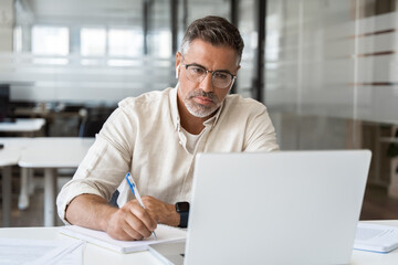 Focused middle-age Hispanic man using laptop computer for business studying, watch online virtual webinar training meeting, video call, make notes. Mature Latin businessman work in office, copy space.