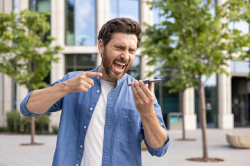 Angry and aggressive young man standing on the city street and talking loudly on the phone through...