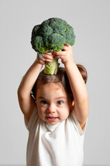 Cute caucasian brunette toddler girl with two tails in white dress having fun holding broccoli at the head, Montessori method on white background 