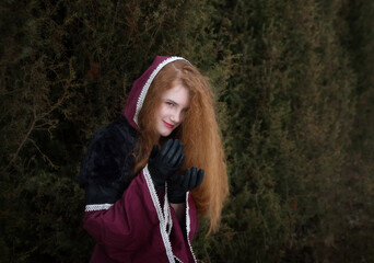 A young girl in an ancient dress with a hood and red hair against the backdrop of a hedge.