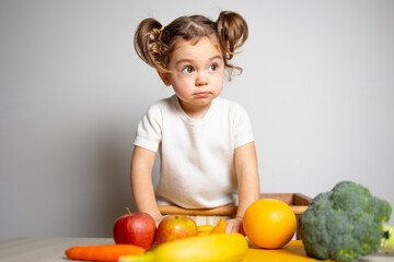 Fototapeta premium Cute caucasian brunette toddler girl with two tails in white dress staying at the learning tower thinking, near vegetables, Montessori method on white background 