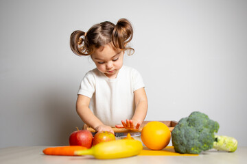 Fototapeta premium Cute caucasian brunette toddler girl with two tails in white dress staying at the learning tower and cutting apple, Montessori method on white background 