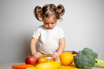 Fototapeta premium Cute caucasian brunette toddler girl with two tails in white dress staying at the learning tower and cutting apple, Montessori method on white background 