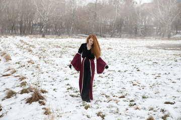 Fototapeta na wymiar A red-haired girl with flowing hair in a beautiful burgundy dress on a spring snow-covered field.