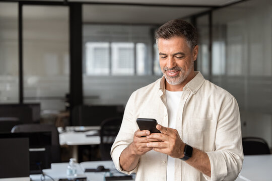 Smiling mature Latin or Indian businessman holding smartphone in office. Middle aged manager using cell phone mobile app. Digital technology application and solutions for business success development.