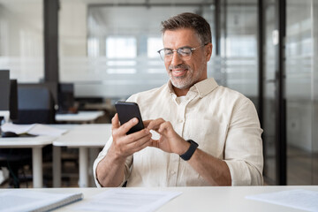 Middle aged Hispanic business manager ceo using cell phone mobile app, laptop. Smiling Latin or...