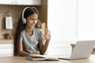 Happy pre teen schoolchild girl using wireless headphones and laptop at home, doing school homework...