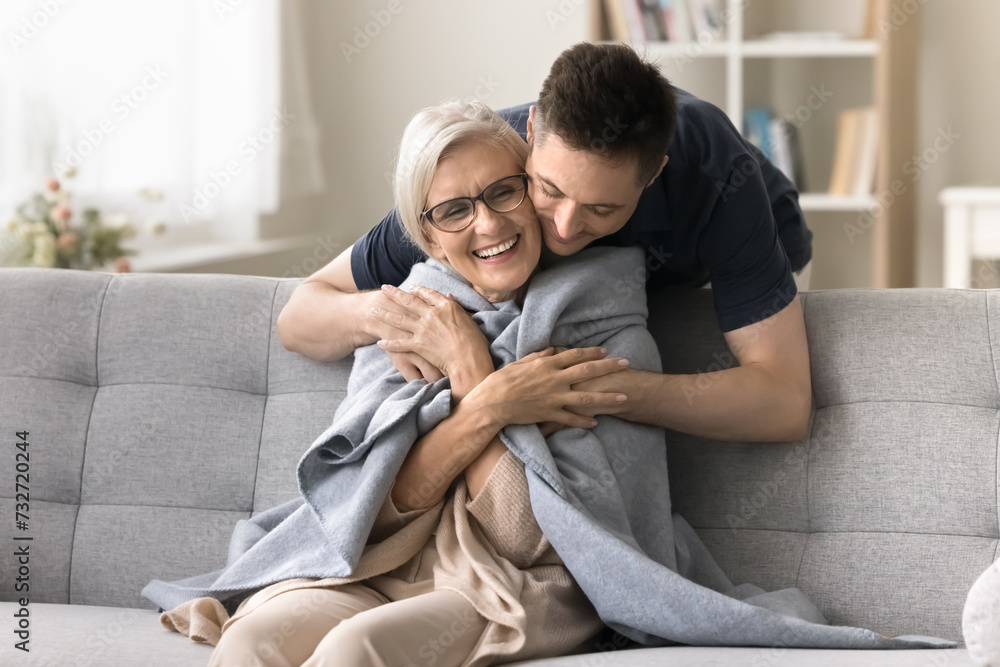 Wall mural Positive young adult son man caring for happy elderly mature mother sitting on comfortable sofa, throwing warm plaid, scarf on moms shoulders, hugging from behind with love, affection