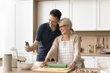 Happy young adult man and senior blonde mom baking in kitchen and talking on video call on smartphone, holding mobile phone, hugging, looking at gadget screen, smiling, laughing