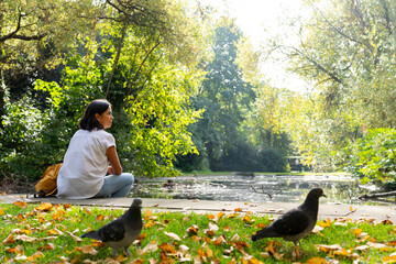 Latin woman sitting enjoying a beautiful view of a pond in a small park in Dublin with two pigeons...