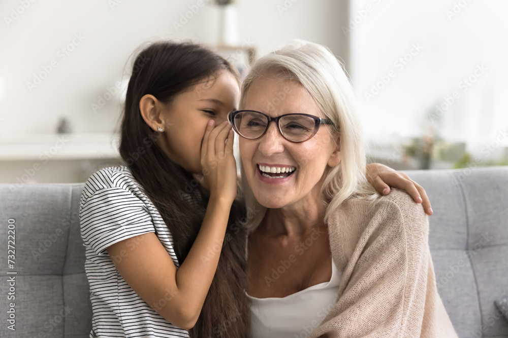 Wall mural pretty adorable granddaughter telling secret to happy laughing grandma, whispering at ear. cheerful 