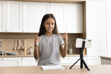 Positive young schoolkid pre teen blogger girl speaking at smartphone fixed on tripod holder. Child talking on learning video call, making preparing online report for class studying project