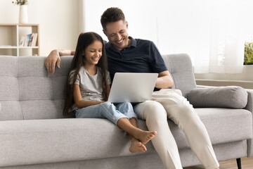Cheerful young dad and pretty little schoolkid girl using Internet web service on laptop on home...