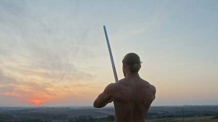 Muscular shirtless man with sword practicing slashing movements in a low angle view against the sunset sky in a side on low angle rear view