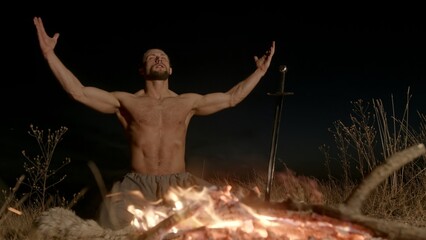 Shirtless male warrior sitting on knees near burning fire with sword and praying to pagan gods at night in nature